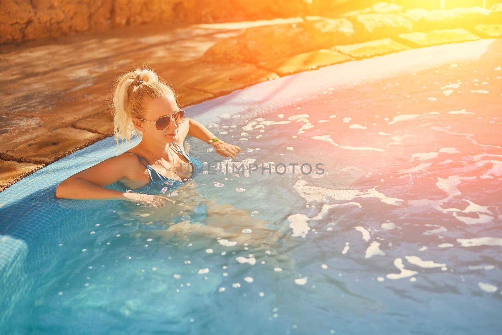 Woman in blue swimsuit and sunglasses relaxing in outdoor pool with clean transparent turquoise water. Woman sunbathing in bikini at tropical resort. Sun flare