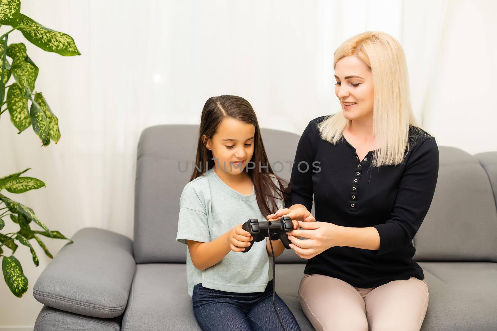 good relationship cute little girl with young mother using joystick playing video game sitting together in living room enjoying family holiday. by Andelov13