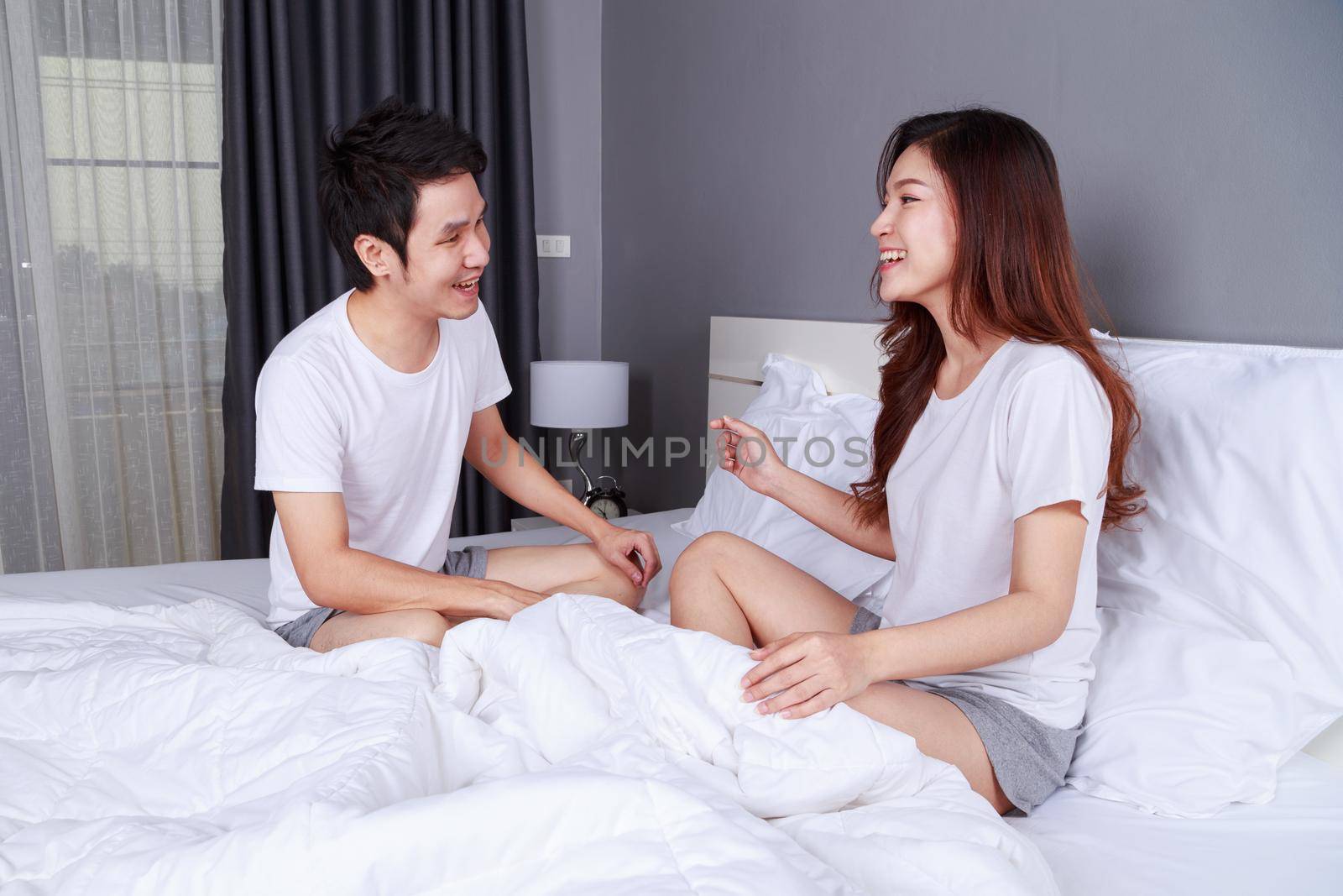 cheerful young couple talking and laughing while sitting on bed in bedroom by geargodz