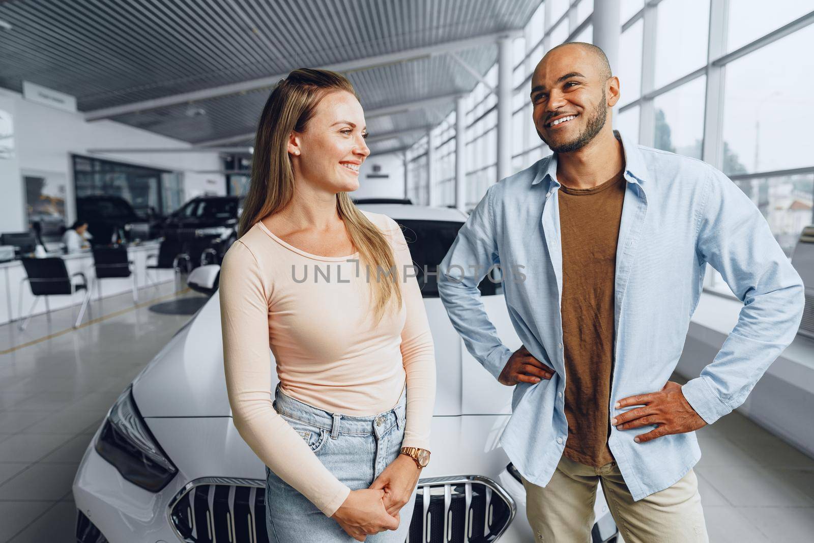 Happy couple of caucasian woman and african american man standing near their new luxury car inside car salon