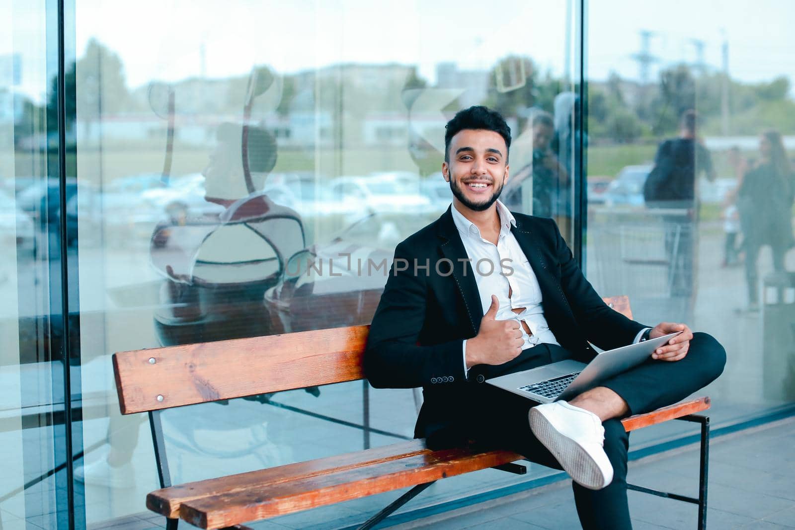 Entrepreneur uses laptop wearing sunglasses. Young handsome businessman arab muslims in business center wearing dressed in black elegant suit on building background.