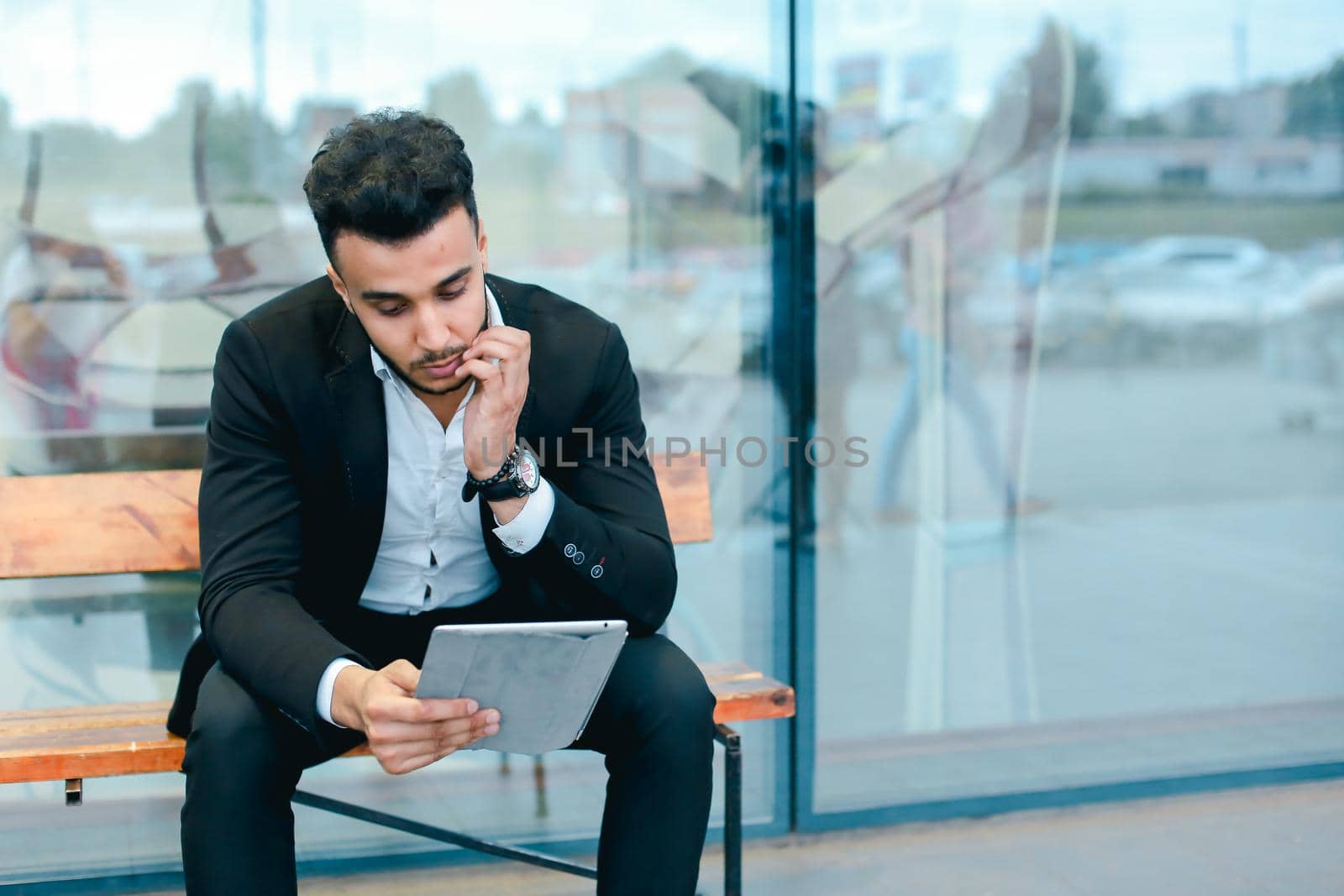 Entrepreneur smiling man puts on sunglasses uses tablet and looks at. Young handsome businessman arab muslims in business center wearing dressed in black elegant suit on building background.