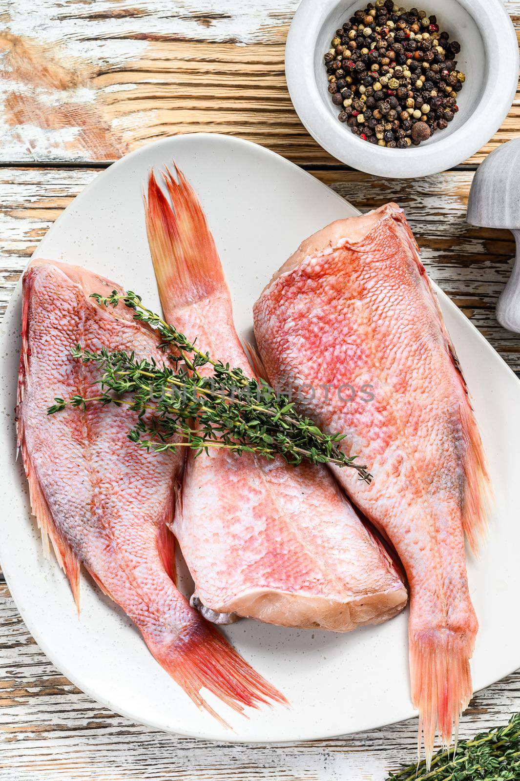 Whole raw red perch or sea bass fish on a plate. White wooden background. Top view.