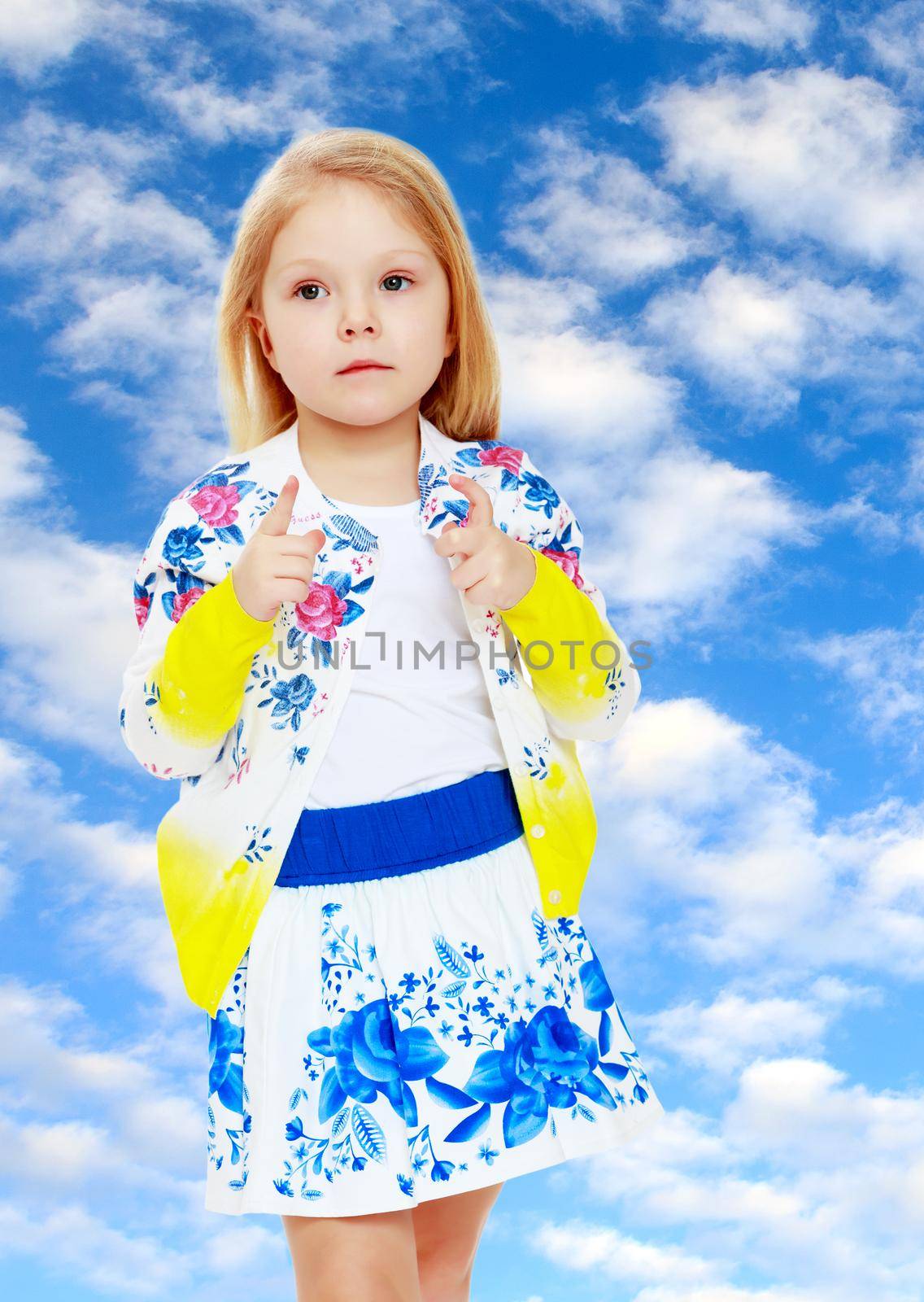 Pretty little blonde girl in a short summer skirt with Russian national ornament.On the background of summer blue sky and fluffy clouds.