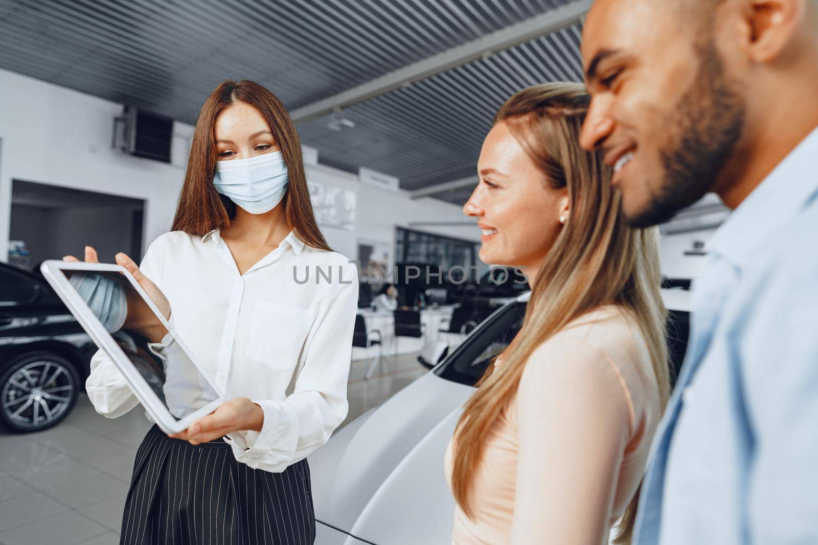 Car saleswoman wearing medical mask shows buyers couple something on digital tablet. .New pandemic job requirements concept