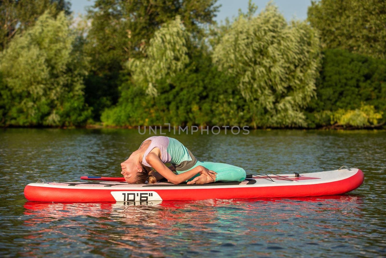 Young woman doing yoga on sup board with paddle. Yoga pose, side view - concept of harmony with the nature, free and healthy living, freelance, remote business.