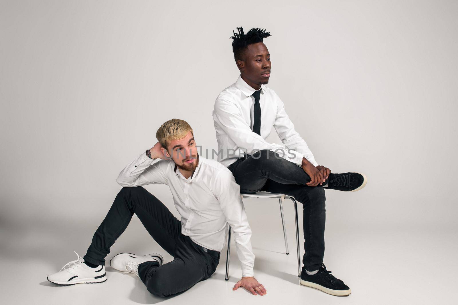 Joyful relaxed african and caucasian boys in white and black office clothes laughing and posing at white studio background with copy space. One man is sitting on the floor, the second is sitting on a chair