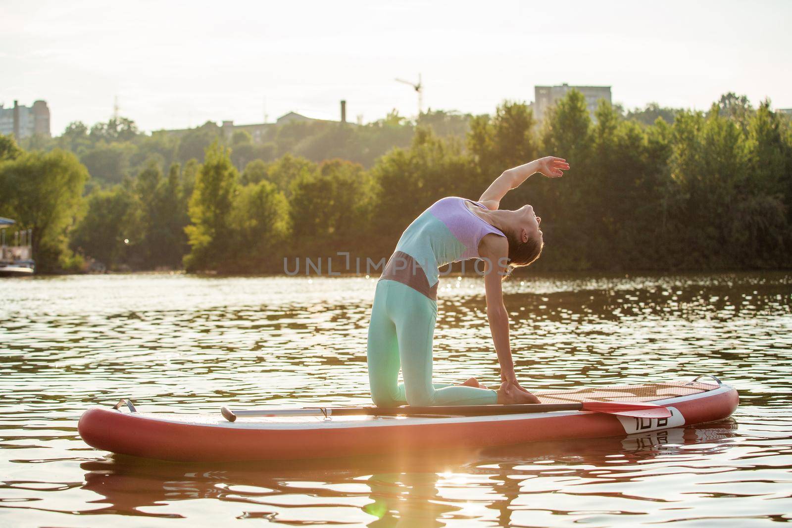 Young woman doing yoga on sup board with paddle. Yoga pose, side view - concept of harmony with the nature, free and healthy living, freelance, remote business.
