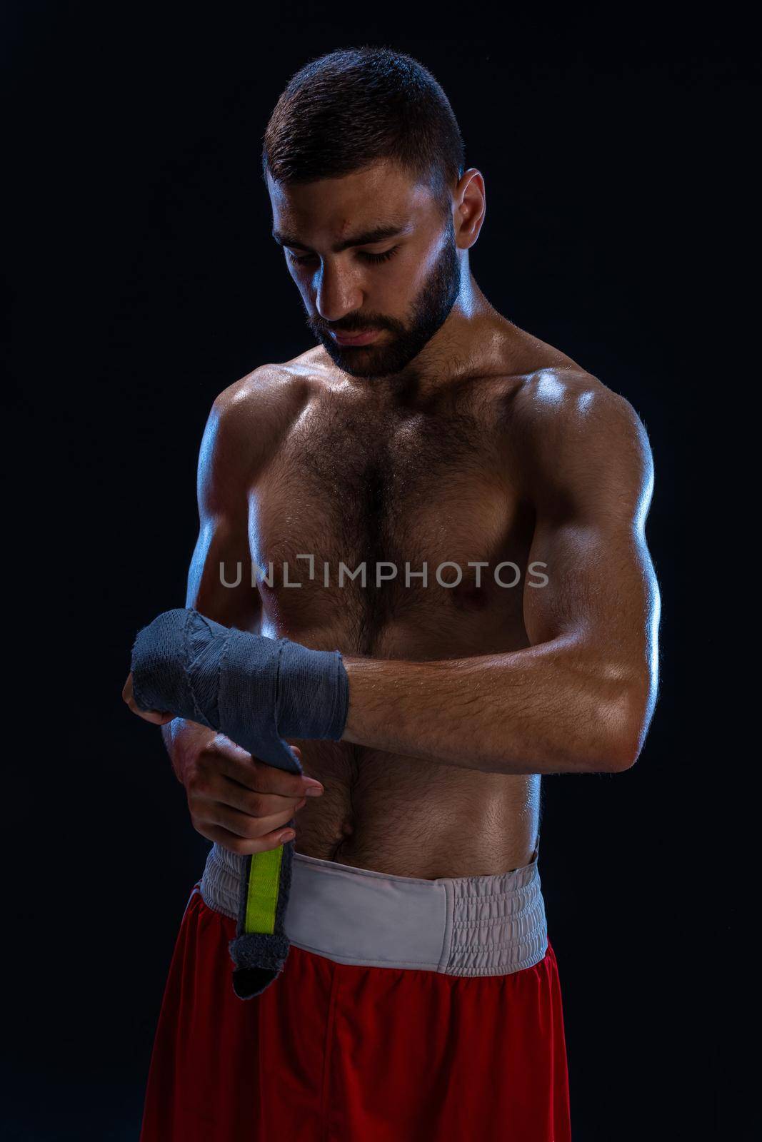 Sports boxer man pulls on the hand wrist wraps. Oriental male model isolated on black background. Athletic man posing in red shorts without shirt