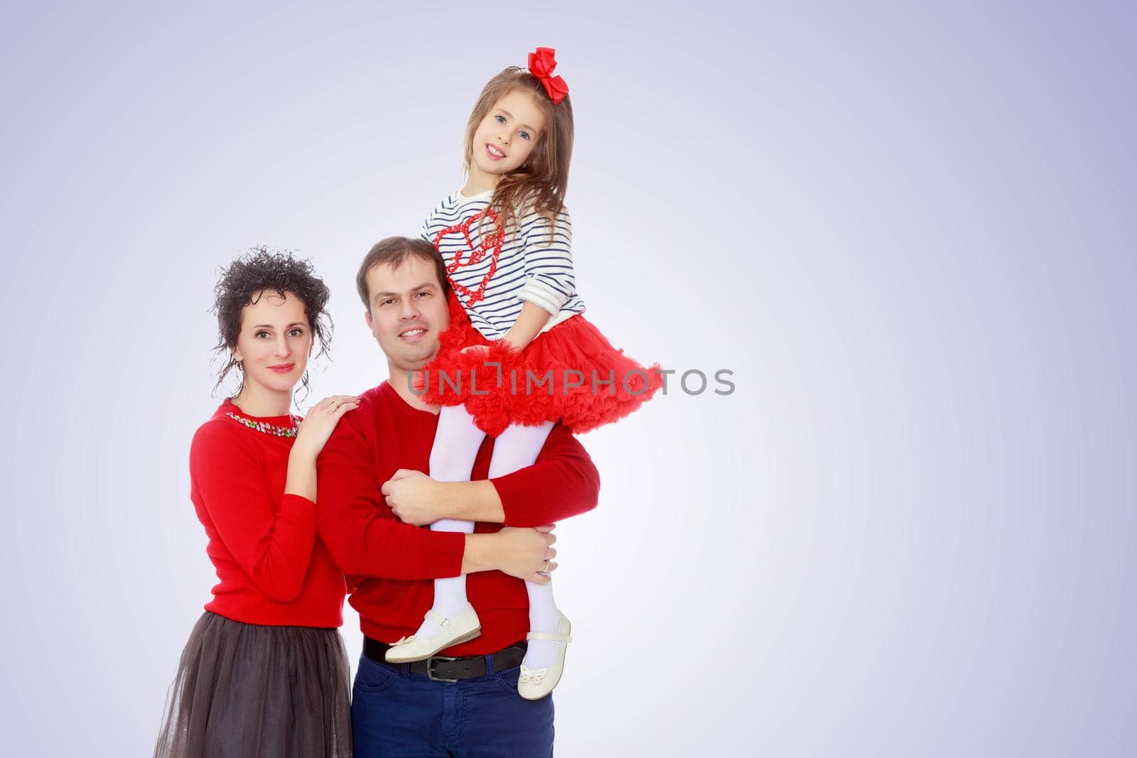 Happy young family dad mom and a little girl in bright red outfits . Dad holds daughter on hands.Purple gradient background.