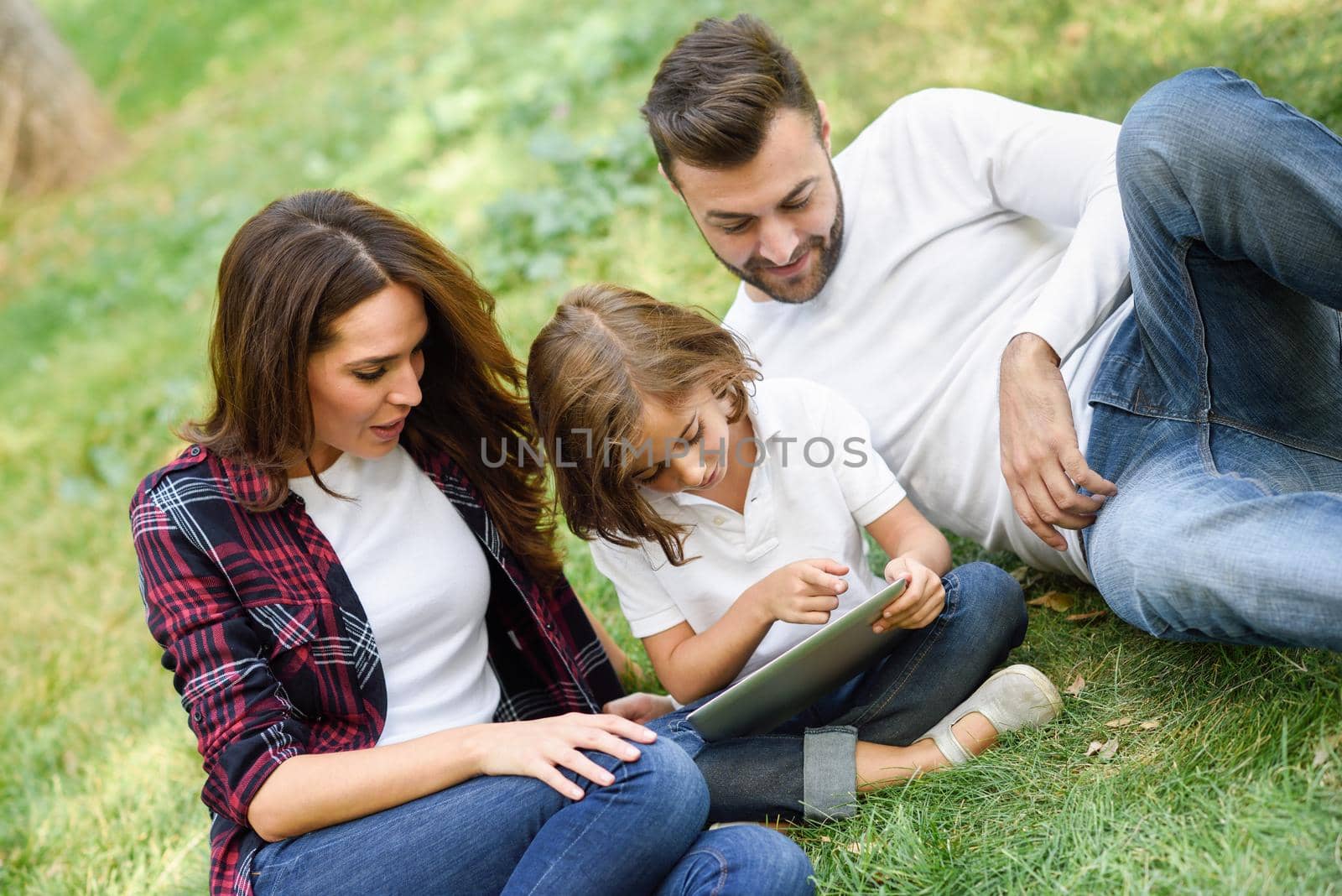 Happy family in a urban park playing with tablet computer. Father, mother and little daughter sitting on the grass laughing.