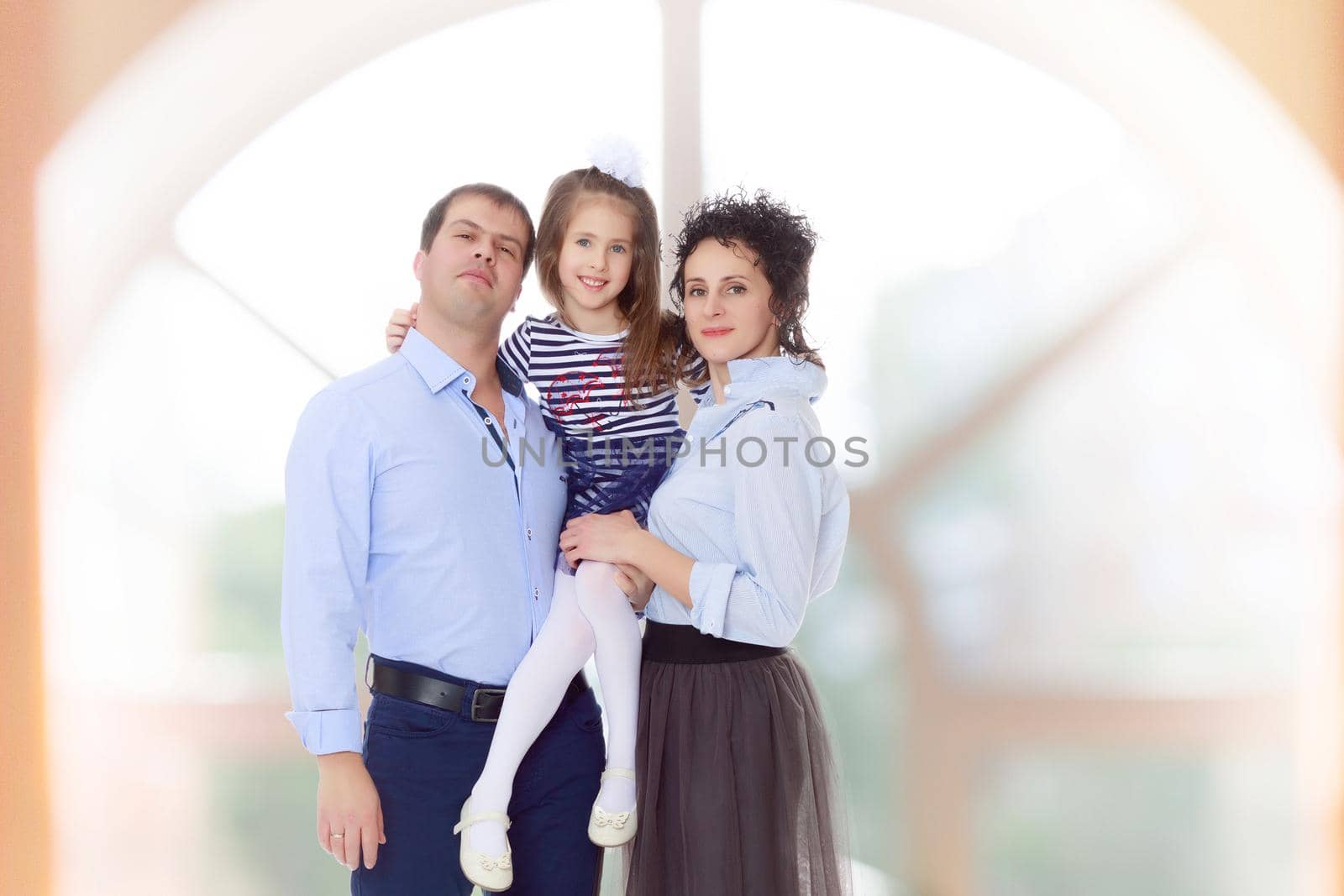 Happy young family, mom dad and little daughter.Parents keep the girl in her arms , and she hugs their neck.In a room with a large semi-circular window.