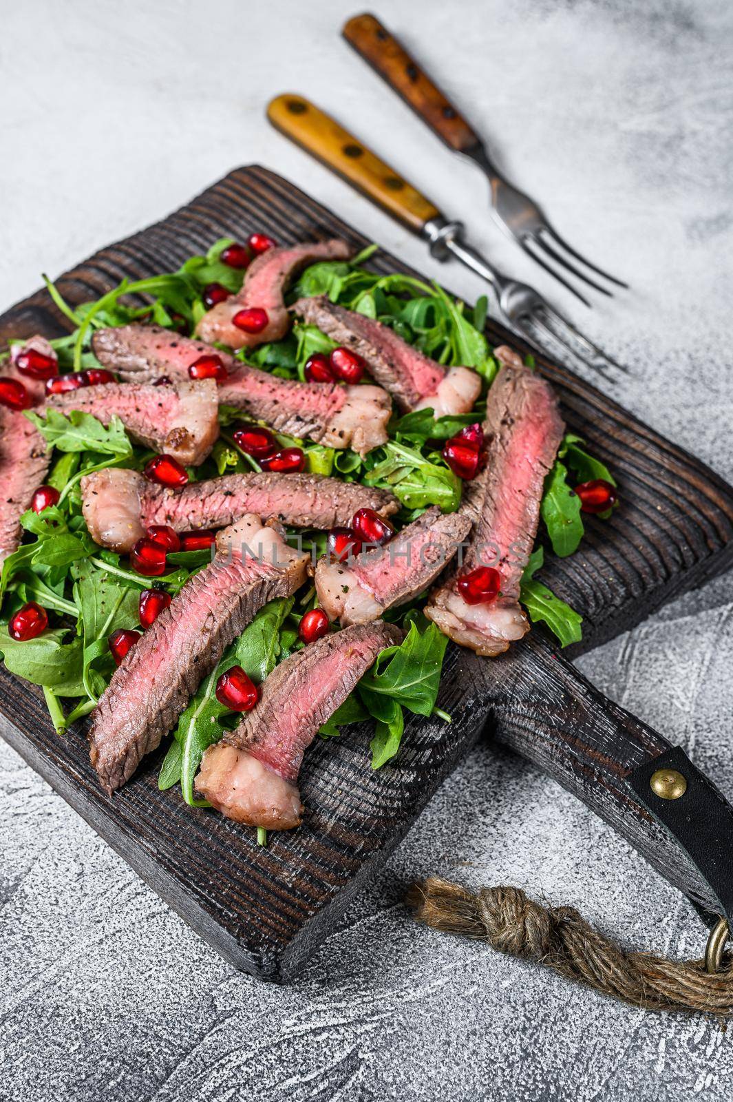 Sliced grilled beef steak with arugula leaves salad on rustic cutting board. White background. Top view.