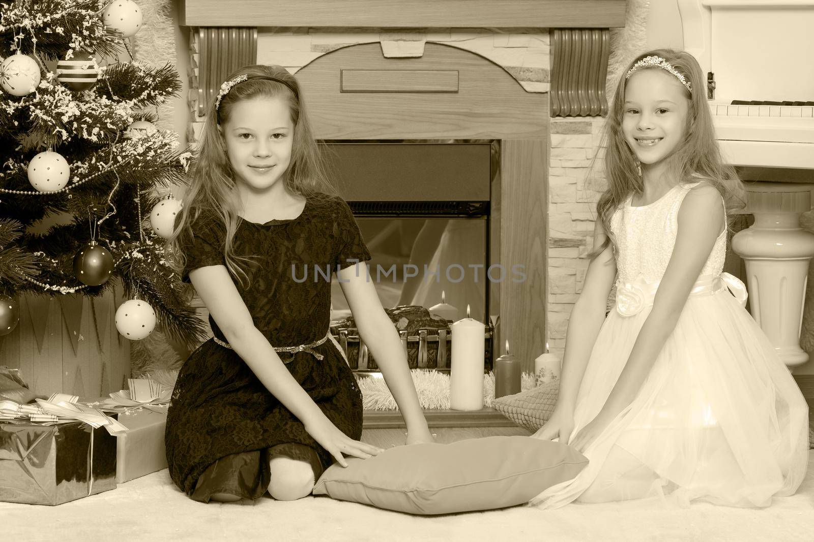 Cute little twin girls, sitting on the floor near the Christmas tree and electric fireplace on which candles are burning.Black-and-white photo. Retro style.