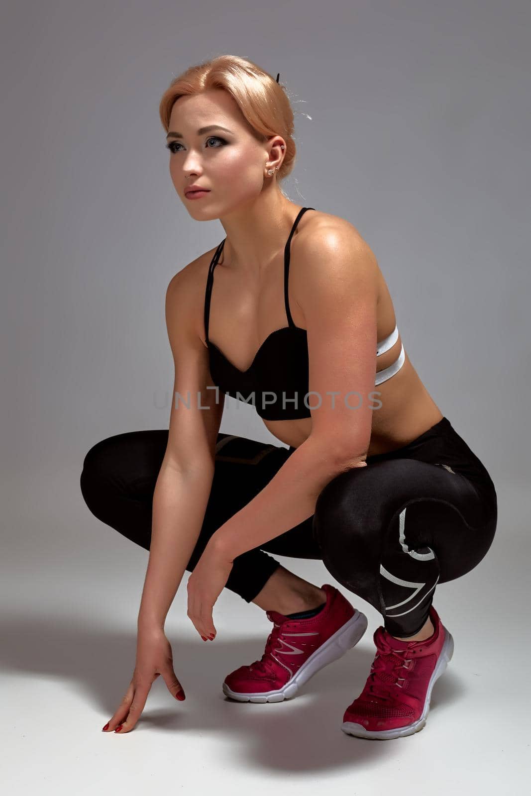 Happy cute young sportswoman sitting over gray background. Studio shot. Young woman wears black leggings and sports top