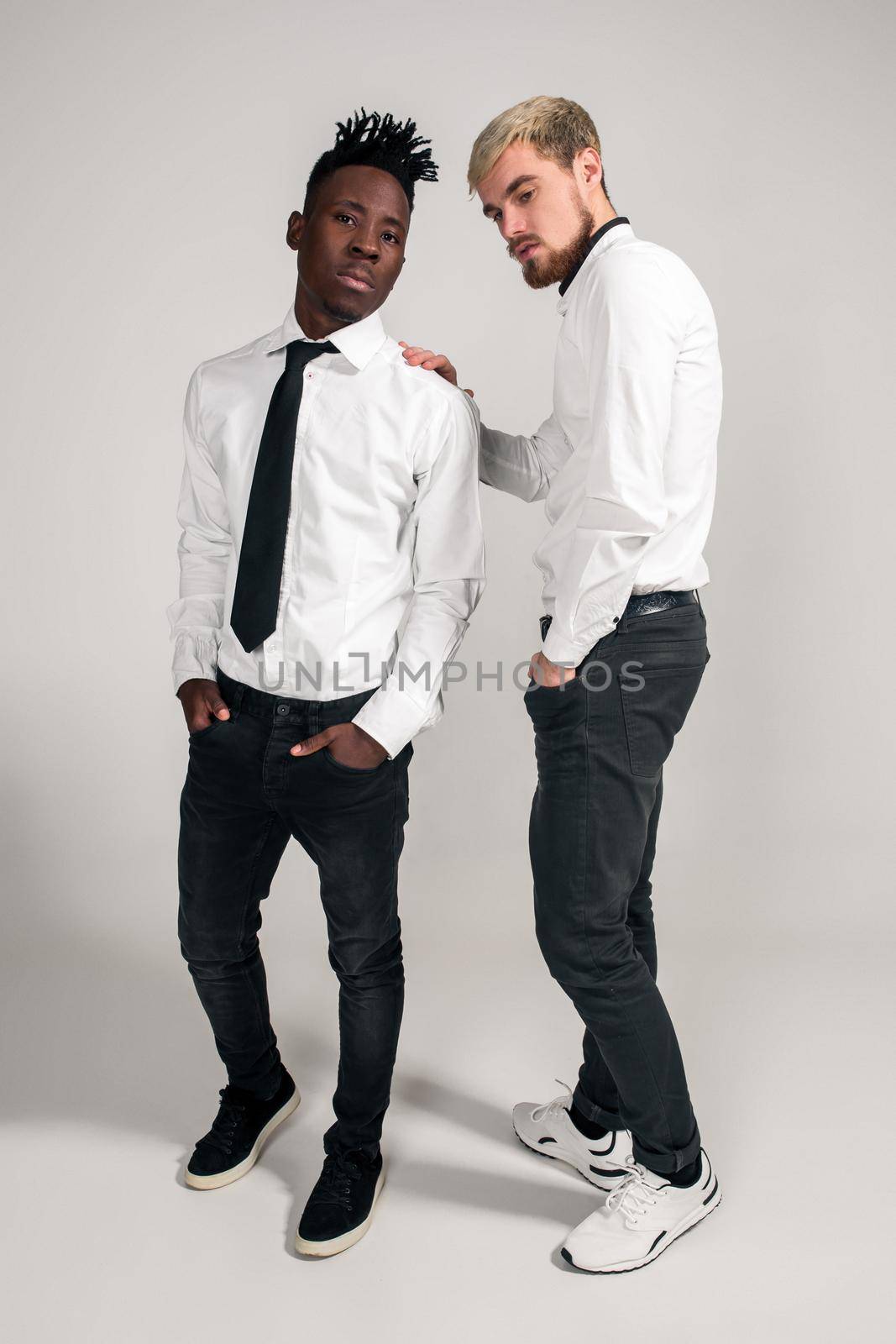 Joyful relaxed african and caucasian boys in white and black office clothes laughing and posing at white studio background with copy space. Full-length photo