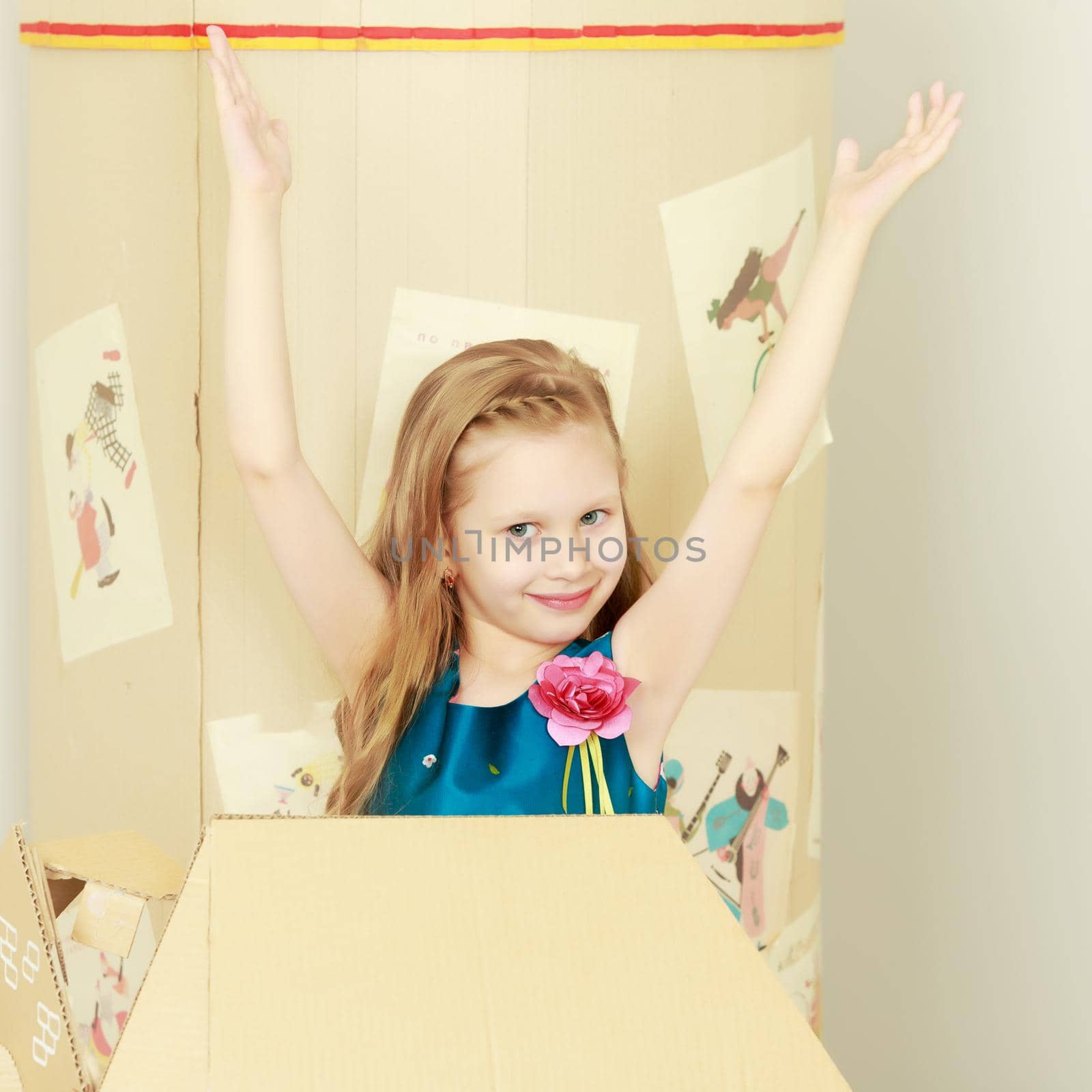 Beautiful little girl with long blond hair in a smart blue dress. Girl playing with a toy wooden car in a cardboard house.