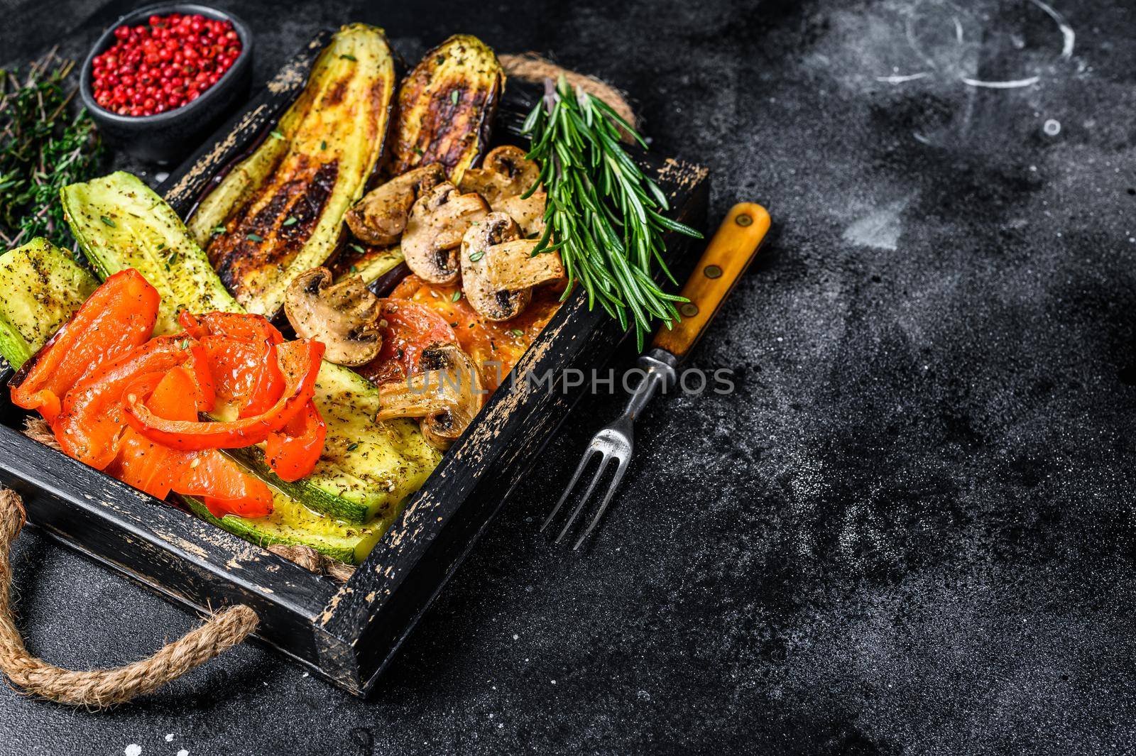 Baked vegetables bell pepper, zucchini, eggplant and tomato in a wooden tray. Black wooden background. Top view. Copy space by Composter