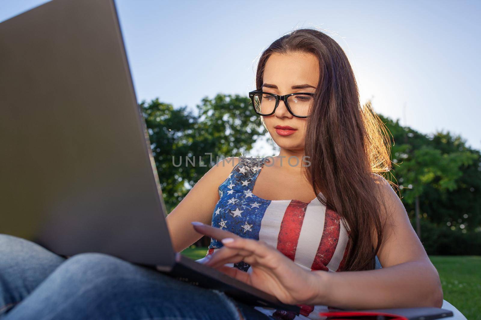 Pretty young woman sitting on bean bag use laptop while resting on grass in park on the sun. Success small business, modern lifestyle, information technology, or online shopping concept