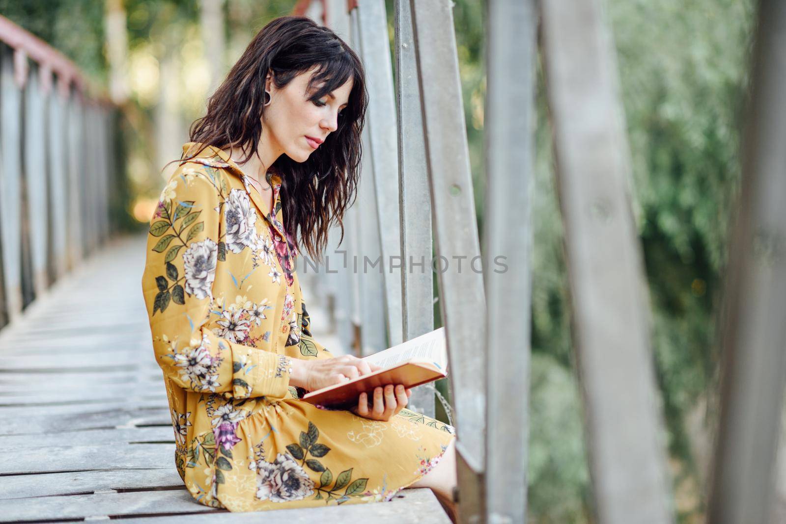 Young woman, wearing flowered dress, reading a book on a rural bridge.