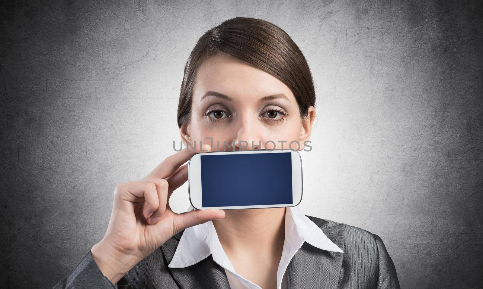 Portrait of young woman covering her mouth with smartphone. Businesswoman showing mobile phone with blank screen. Corporate businessperson on grey wall background. Mobile communication layout.