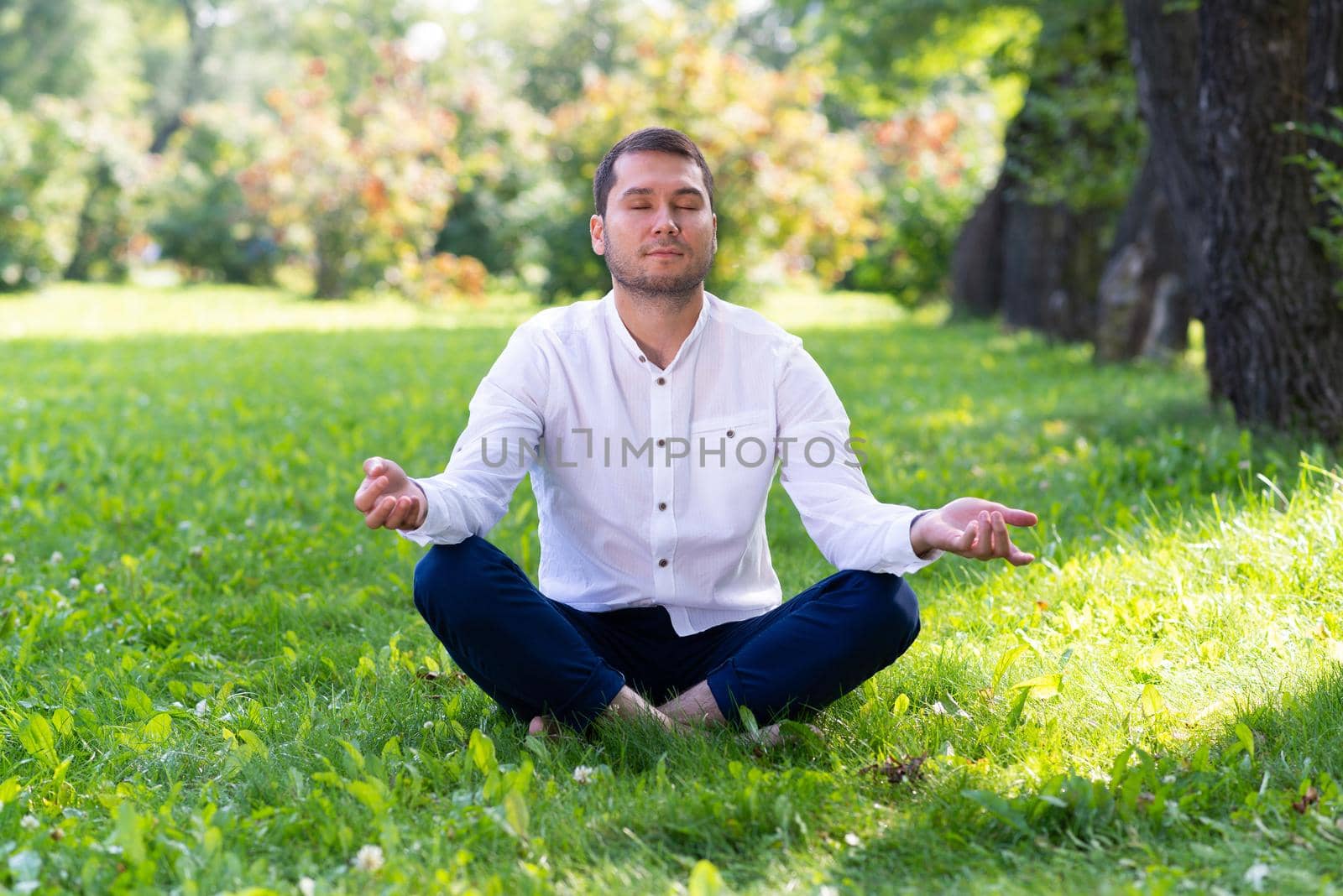 Young man meditates in lotus pose on green grass. Handsome man in casual wear practicing of yoga with closed eyes. Training and meditation outdoor at summer day. Healthy lifestyle and relaxation.