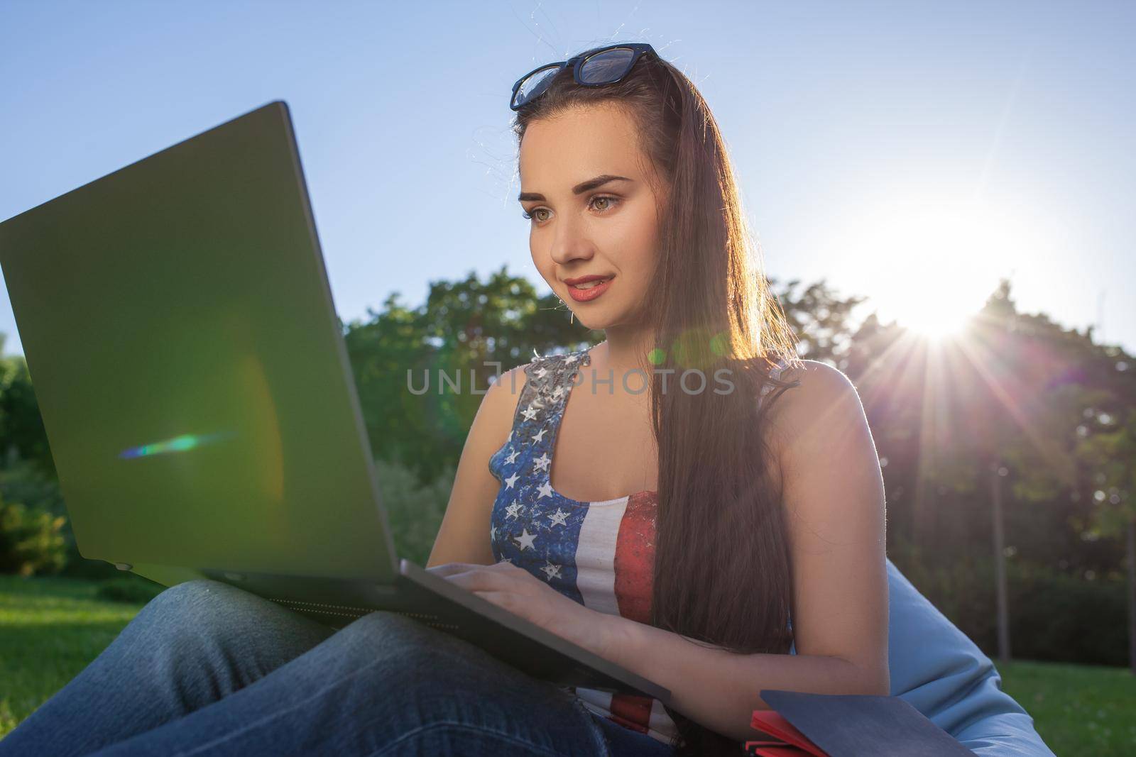 Pretty young woman sitting on bean bag use laptop while resting on grass in park on the sun by nazarovsergey