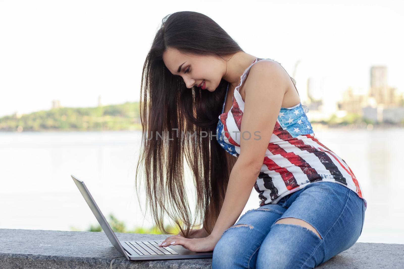 The beautiful young woman sits in the park near river with the laptop. Success small business, modern lifestyle, information technology, or online shopping concept