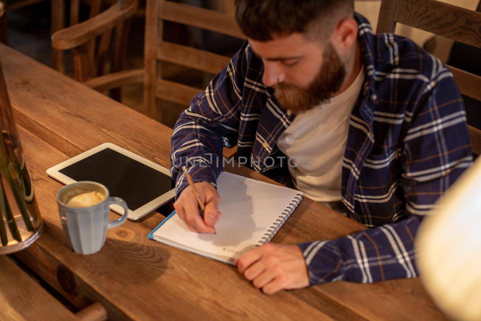 Young bearded businessman sits in cafe, home at table and writes in notebook, near lies tablet computer with black screen. Man is working, studying. Online education, marketing, training.