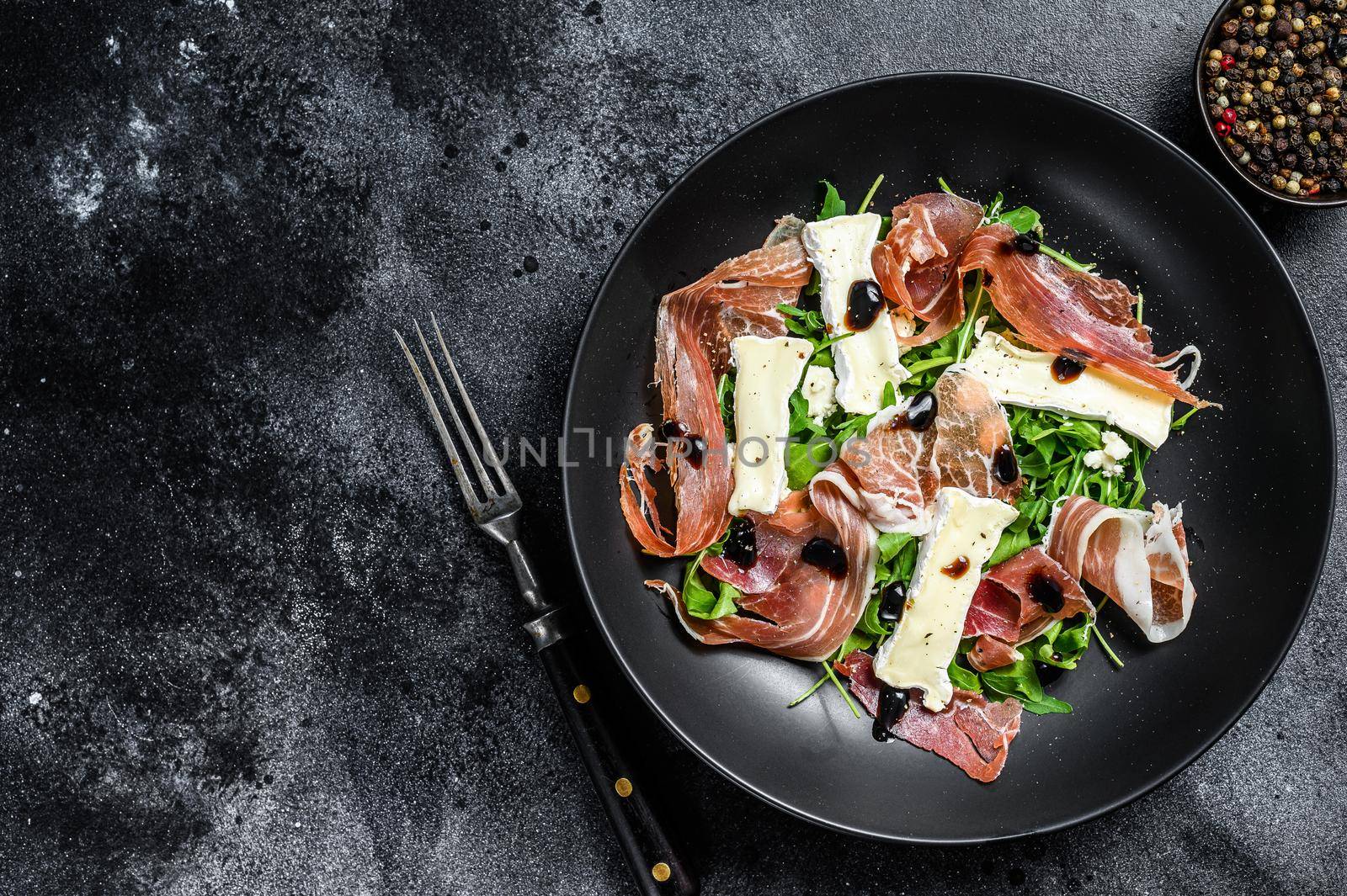 Prosciutto crudo ham salad with brie camembert cheese and arugula on a plate. Black background. Top view. Copy space by Composter
