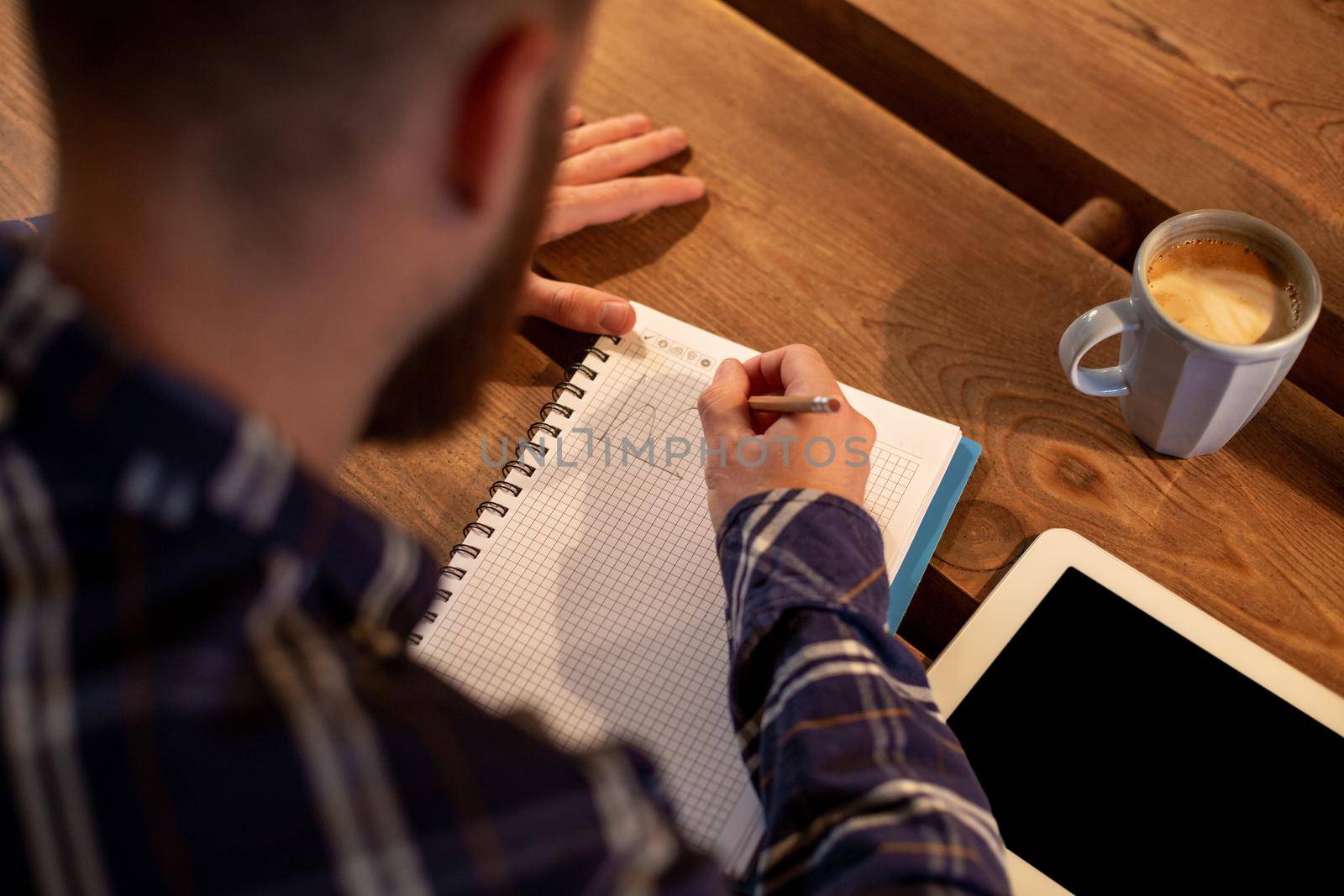 Young bearded businessman sits in cafe, home at table and writes in notebook, near lies tablet computer with black screen. Man is working, studying. Online education, marketing, training.