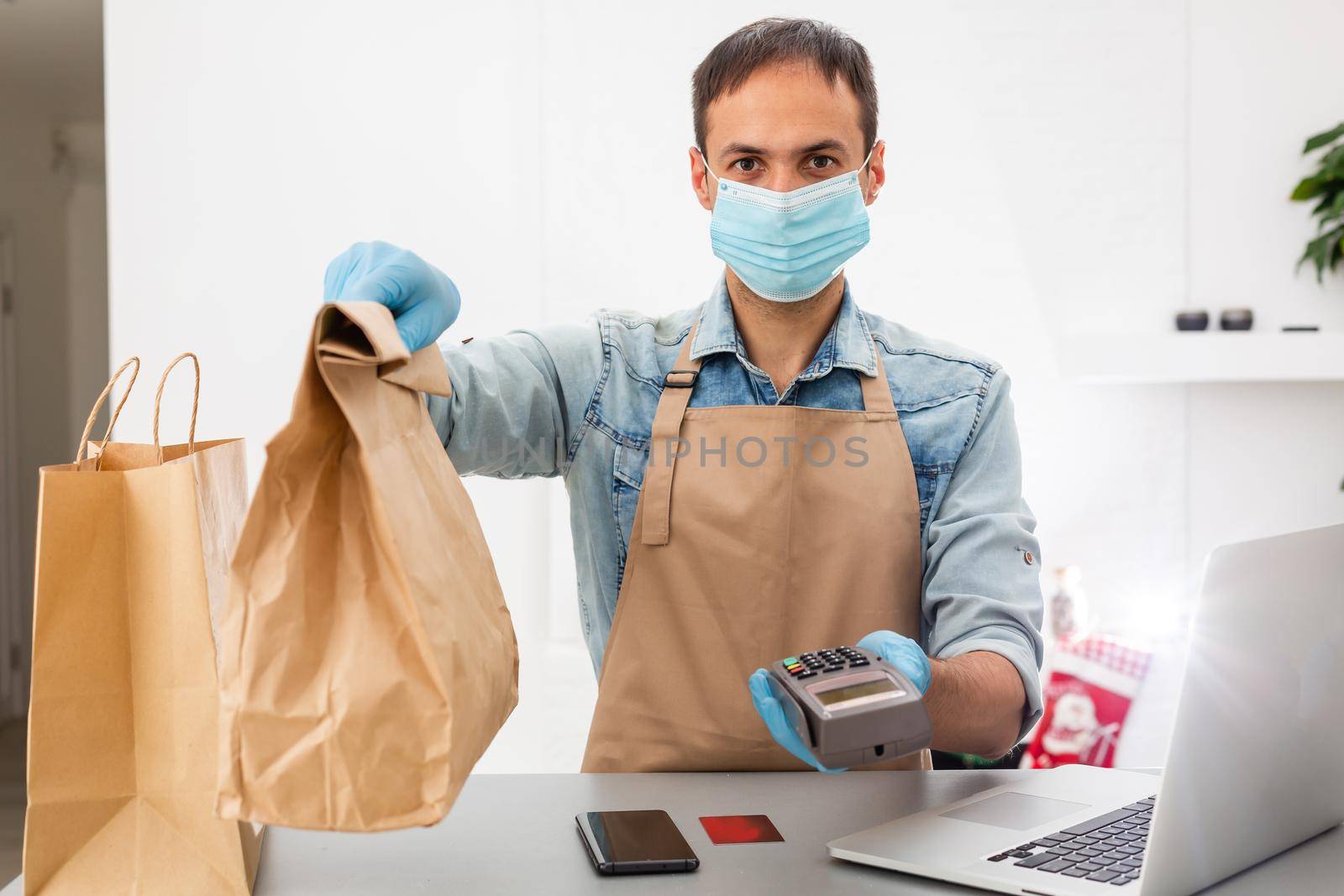 Cashier hand holding credit card reader machine and wearing disposable gloves, paying with smartphone during Covid-19 pandemic.