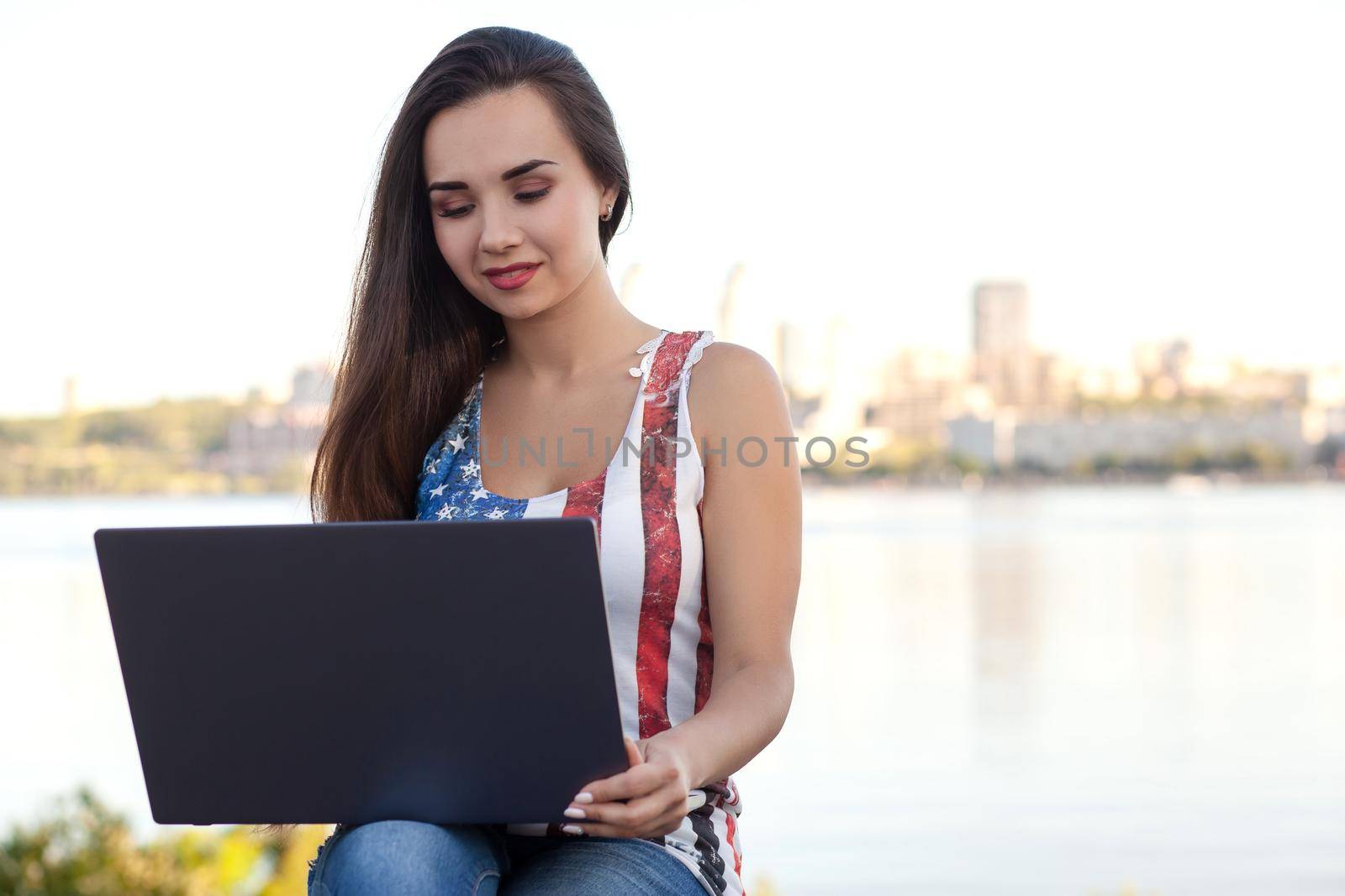 The beautiful young woman sits in the park near river with the laptop. Success small business, modern lifestyle, information technology, or online shopping concept