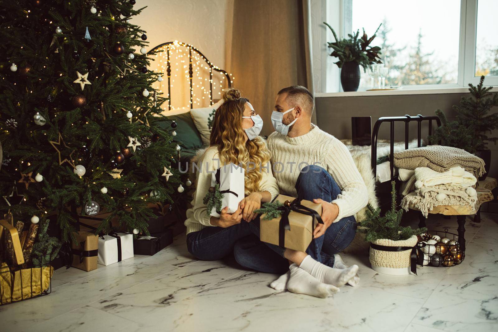 Young man and woman in masks looking each other while holding Christmas gifts in their bedroom