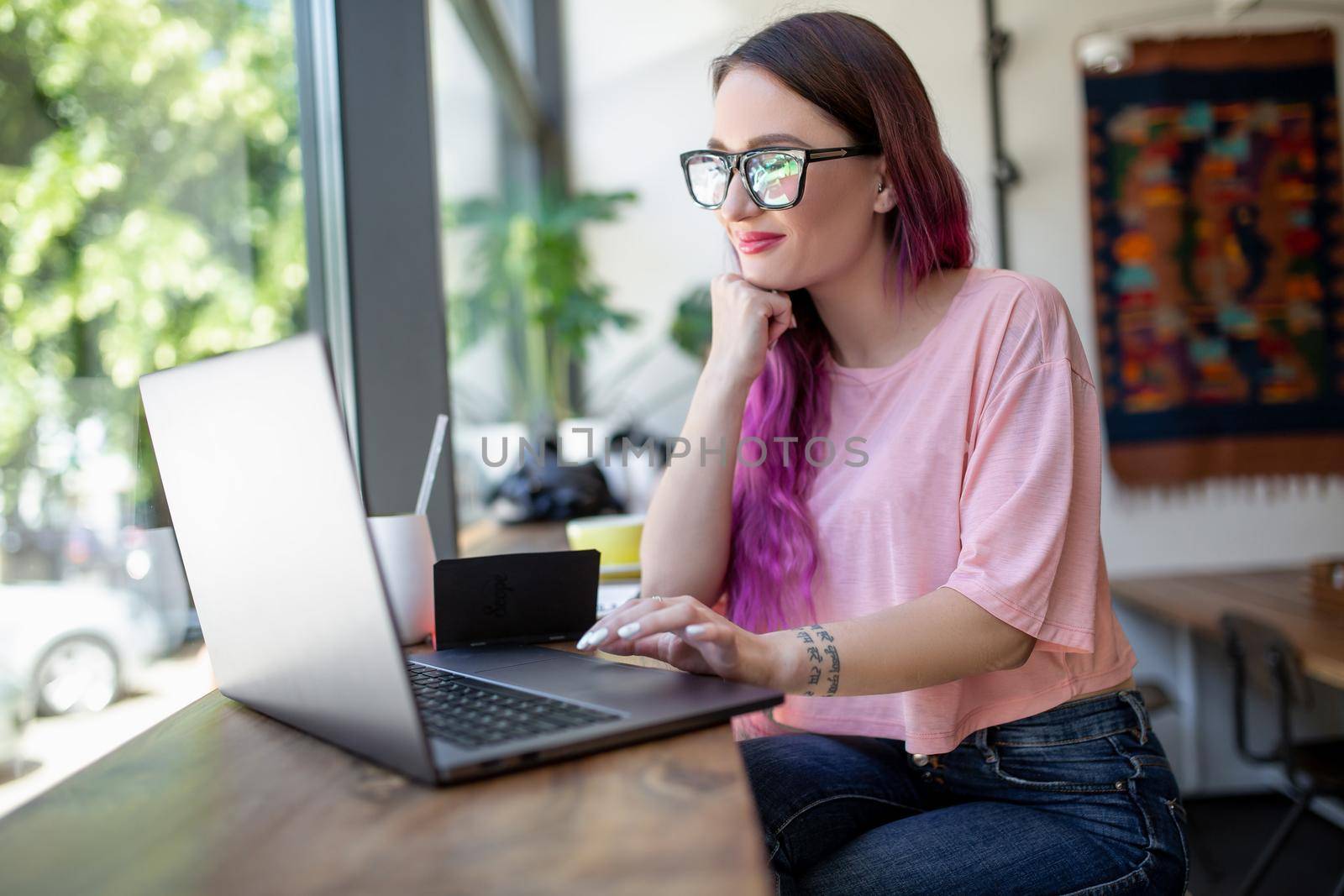 Side view of young businesswoman sitting at table in coffee shop. On table cup of coffee and laptop. In background white wall and window. Girl shopping online.