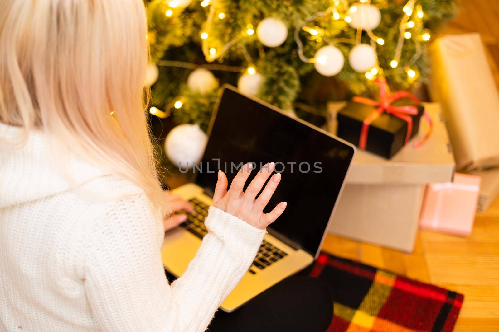 Pretty Young Woman in Trendy Winter Fashion Sitting on the Floor with Crossed Legs Holding Laptop Gadget in Front Christmas Tree Decoration, While Looking at the Camera.