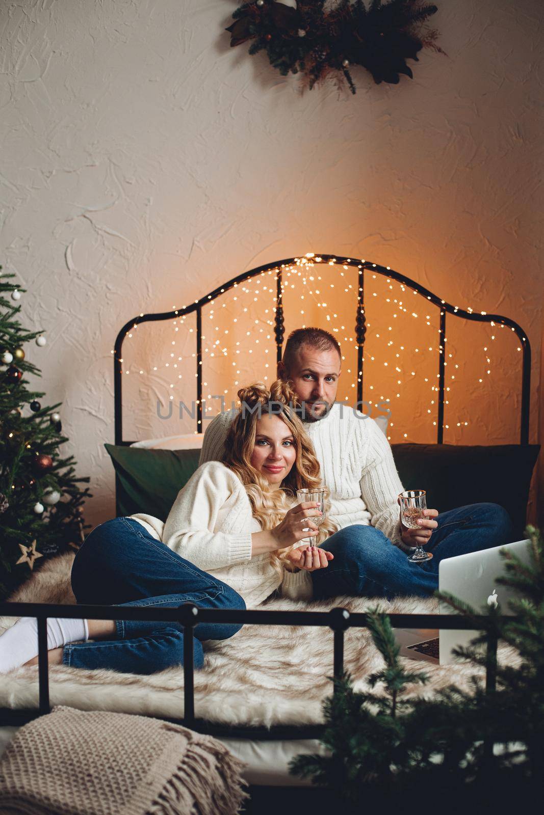 Couple in sweaters and jeans celebrating New Year with drinks and laptop on bed. Lockdown concept.