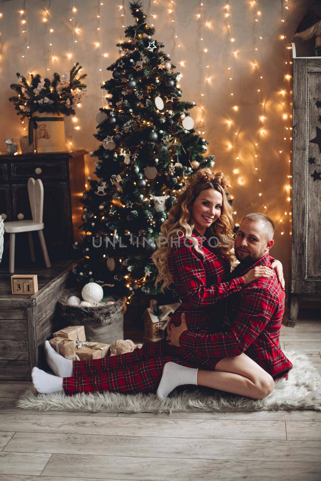 Happy Caucasian couple in checked pajamas sitting embraced by the Christmas tree. Xmas holiday.