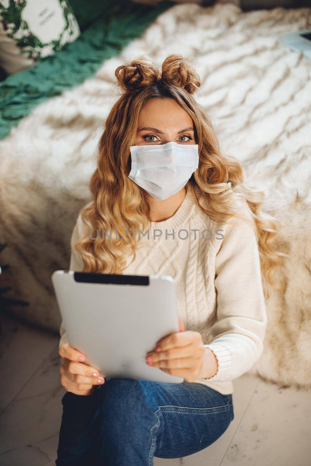 Pretty blonde woman in white sweater in facial mask holding a tablet sitting by bed at xmas.