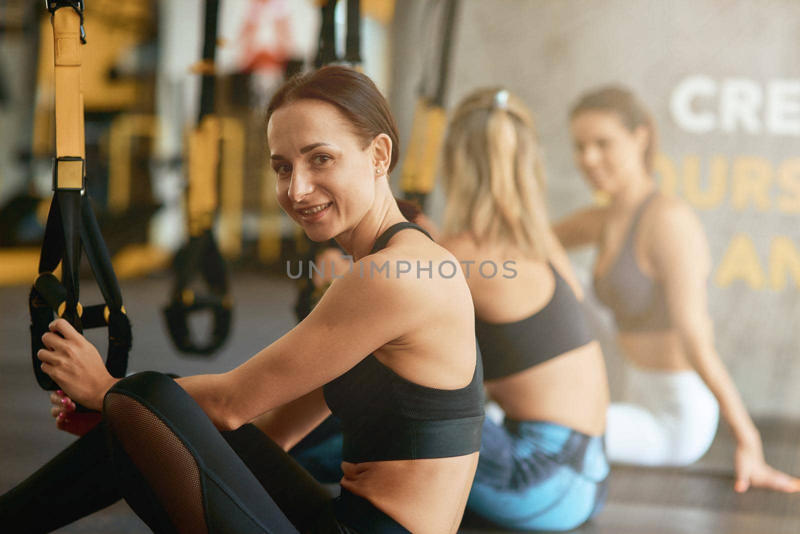 Young happy fitness woman sitting on yoga mat and smiling at camera while having trx training at gym, selective focus. Sport, wellness and healthy lifestyle