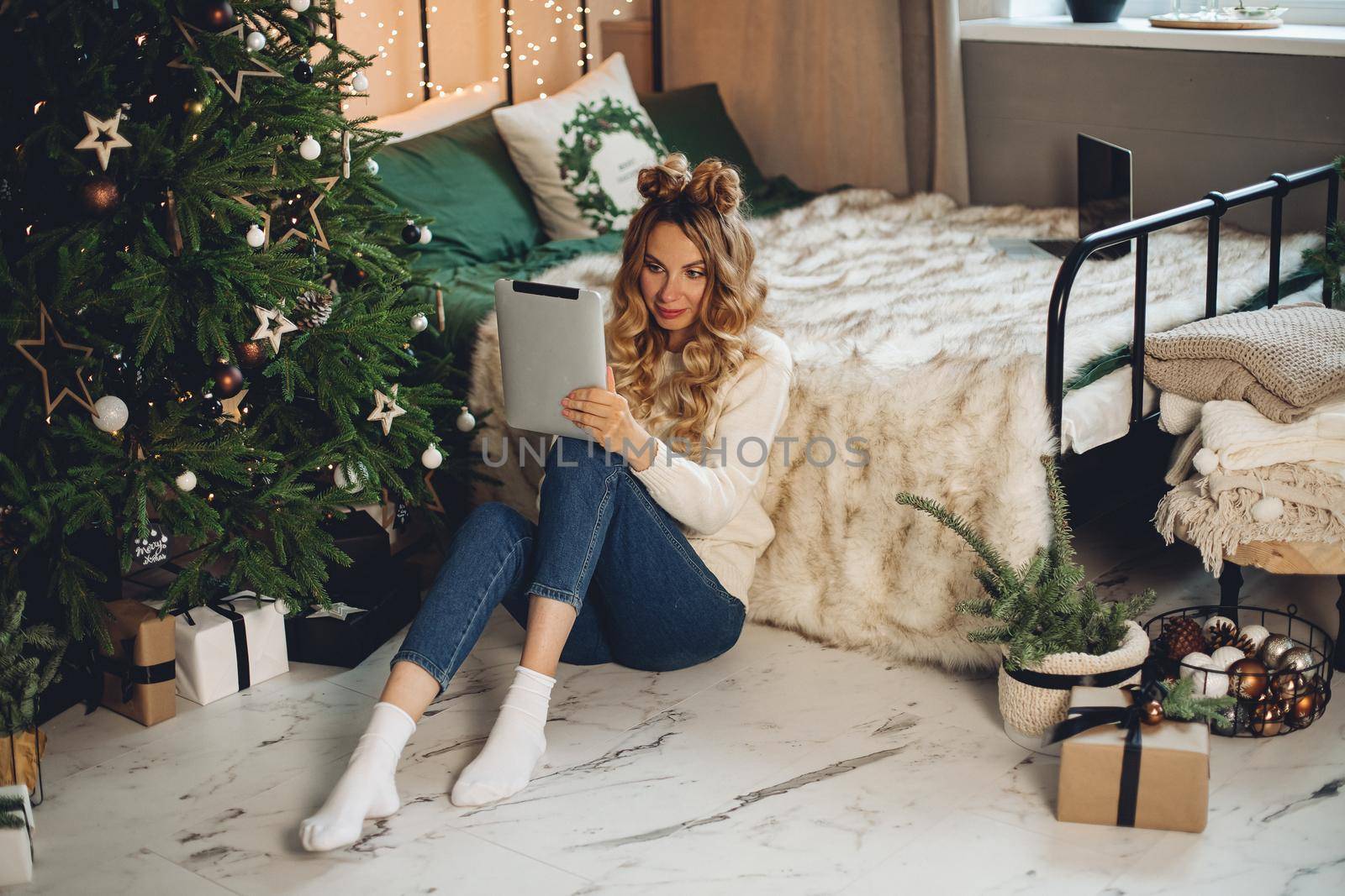 Pretty young woman sitting near Christmas tree and communicating while using digital tablet