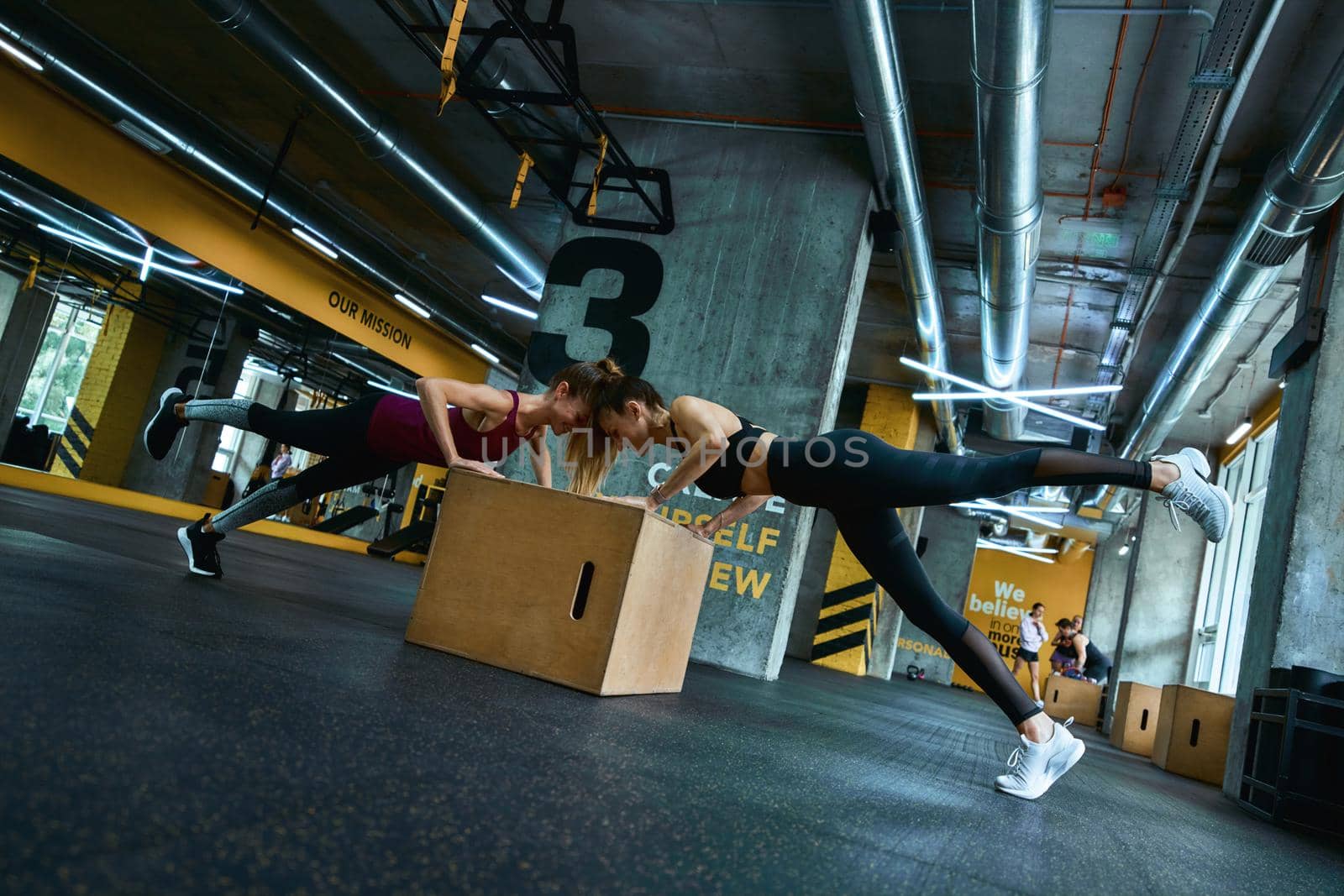 Full length of two young athletic women in sportswear doing push ups on wooden crossfit jump box at gym, exercising together. Sportive people, healthy lifestyle and workout concept