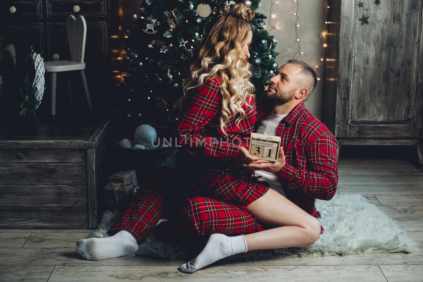 Beautiful couple in red checked pajamas holding wooden calendar with 31st date. New Year’s Eve.