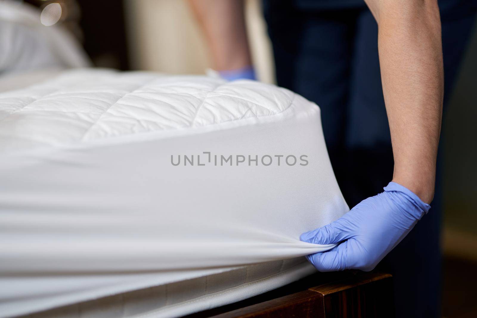 Close up of woman in protective gloves changing clean sheet on white mattress in hotel room. Hotel service concept