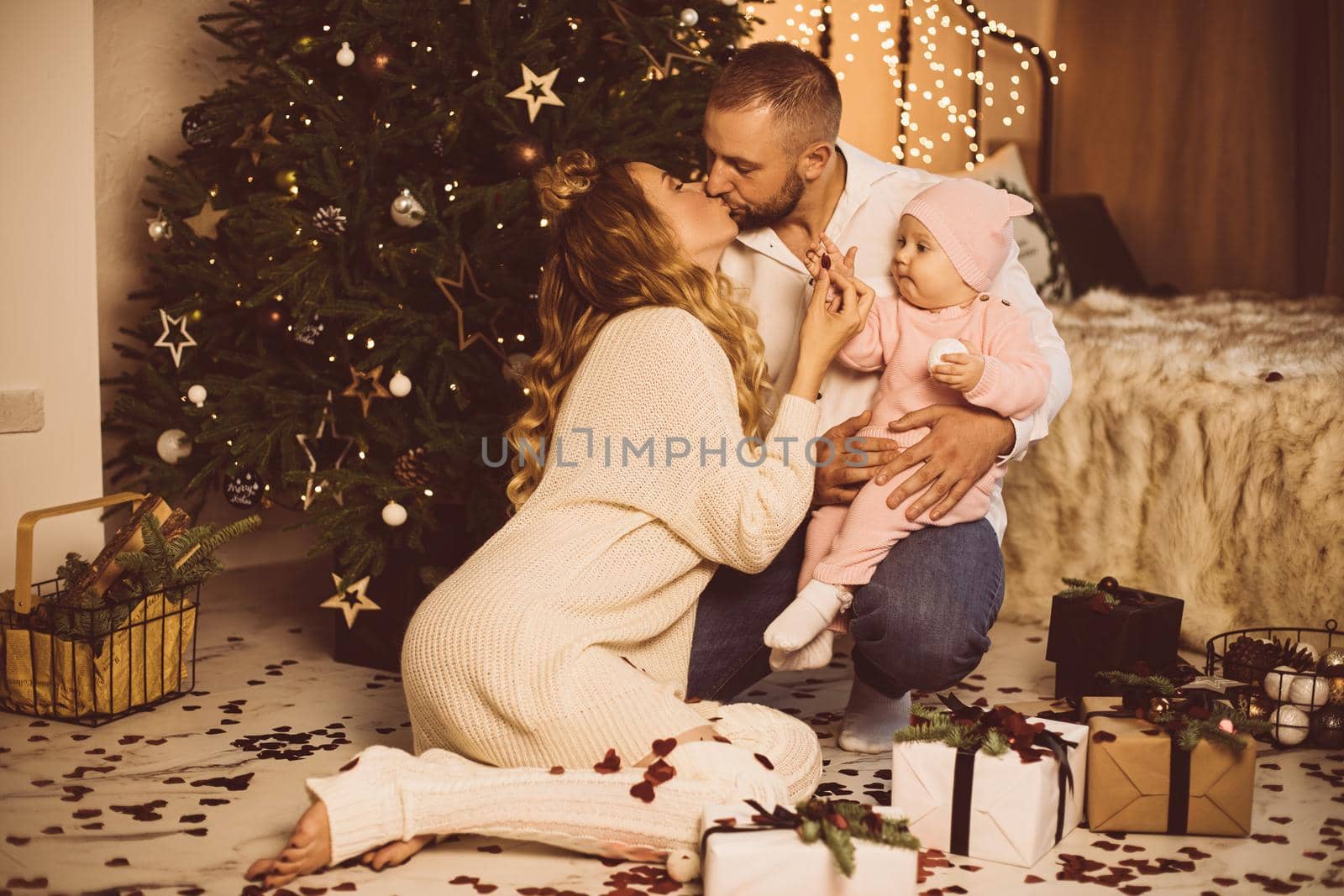 Portrait of kissing parents holding baby daughter playing with confetti in father’s arms. Christmas.