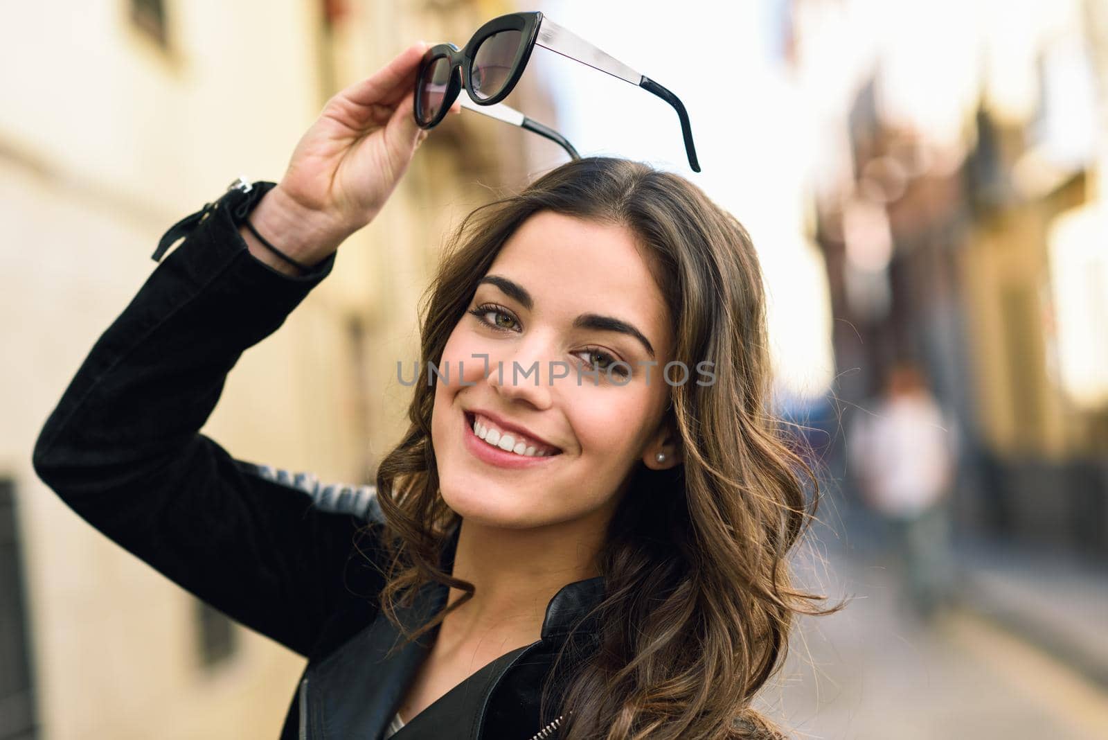 Portrait of young woman smiling in urban background wearing casual clothes with modern sunglasses. Girl with long curly hair
