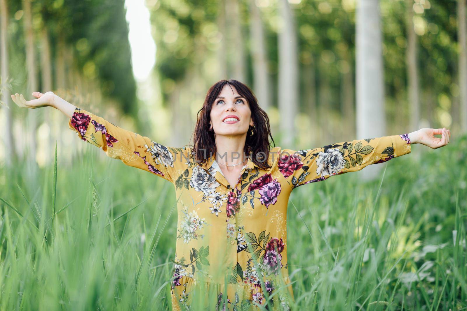 Enjoying the nature. Young woman arms raised enjoying the fresh air in green forest. Female wearing flowered dress.