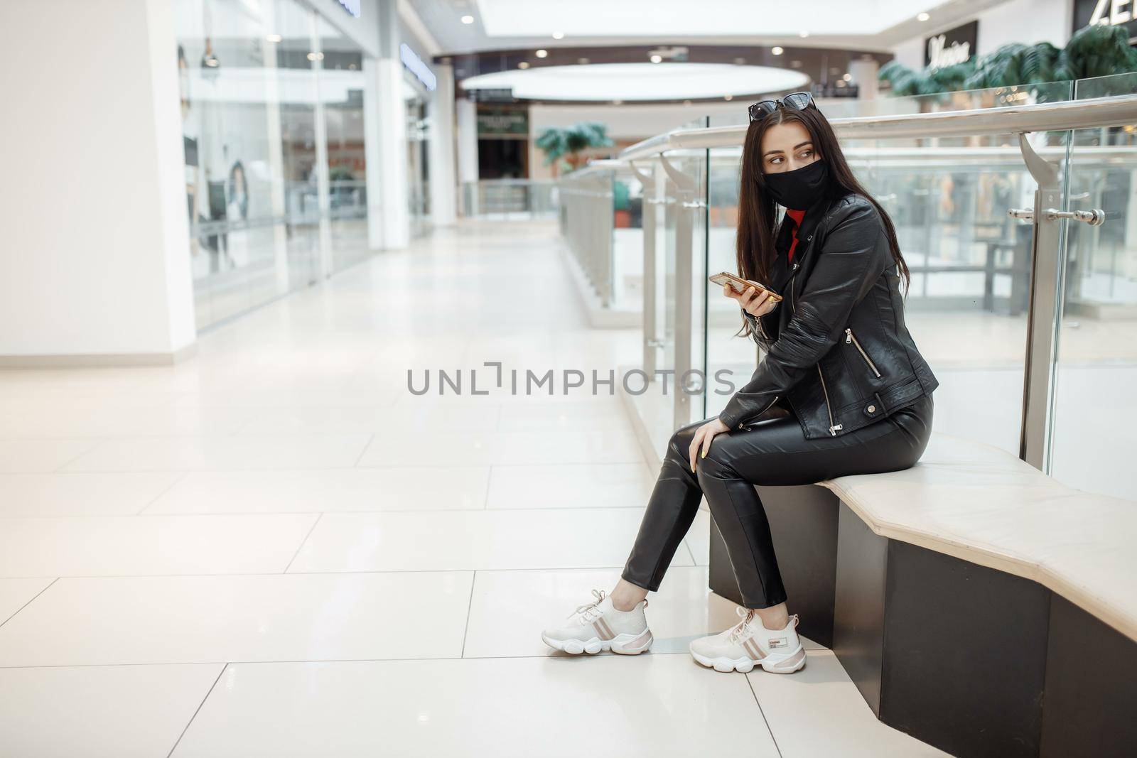 Girl with medical black mask and mobile phone in a shopping center. Coronavirus pandemic. A woman with a mask is standing in a shopping center. A girl in a protective mask is shopping at the mall by UcheaD