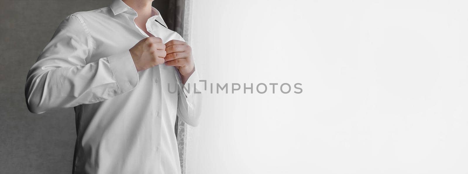 A man clasps buttons on a white shirt with his hands in the light from the window by AYDO8