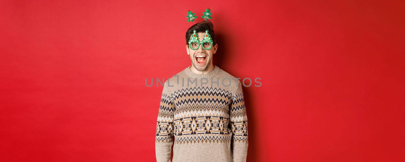 Portrait of surprised and excited handsome guy in party glasses and winter sweater, celebrating christmas and having fun, standing against red background by Benzoix